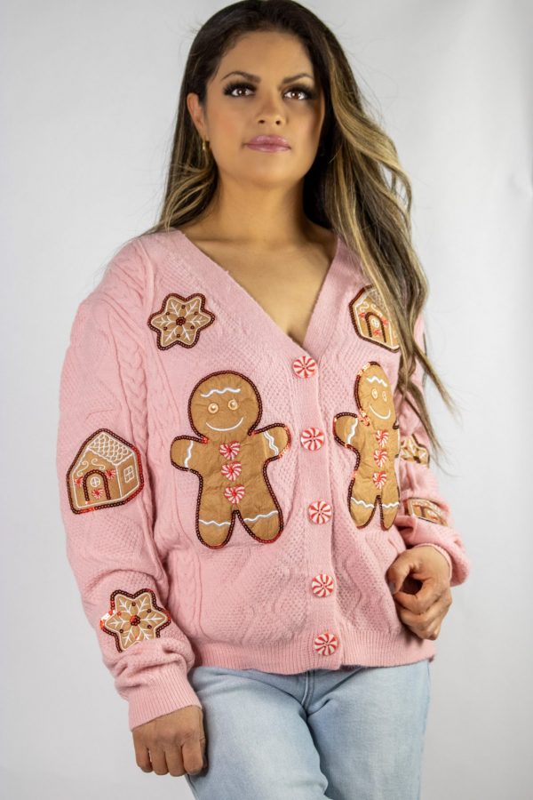 A soft pink knit Gingerbread Man cardigan adorned with playful gingerbread man accents, styled casually with a white tee and jeans. The cozy, festive look is set against a holiday backdrop featuring twinkling lights, pinecones, and a cup of hot cocoa.