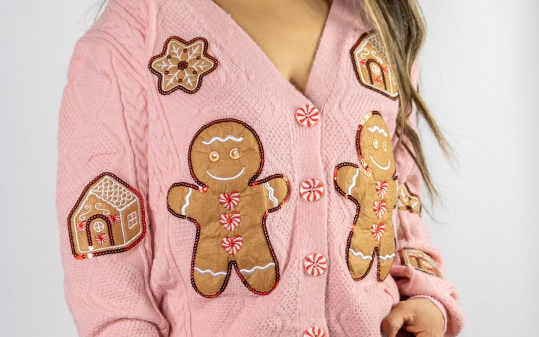 A soft pink knit Gingerbread Man cardigan adorned with playful gingerbread man accents, styled casually with a white tee and jeans. The cozy, festive look is set against a holiday backdrop featuring twinkling lights, pinecones, and a cup of hot cocoa.