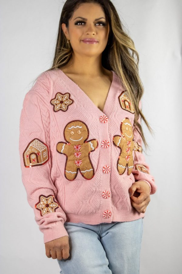 A soft pink knit Gingerbread Man cardigan adorned with playful gingerbread man accents, styled casually with a white tee and jeans. The cozy, festive look is set against a holiday backdrop featuring twinkling lights, pinecones, and a cup of hot cocoa.