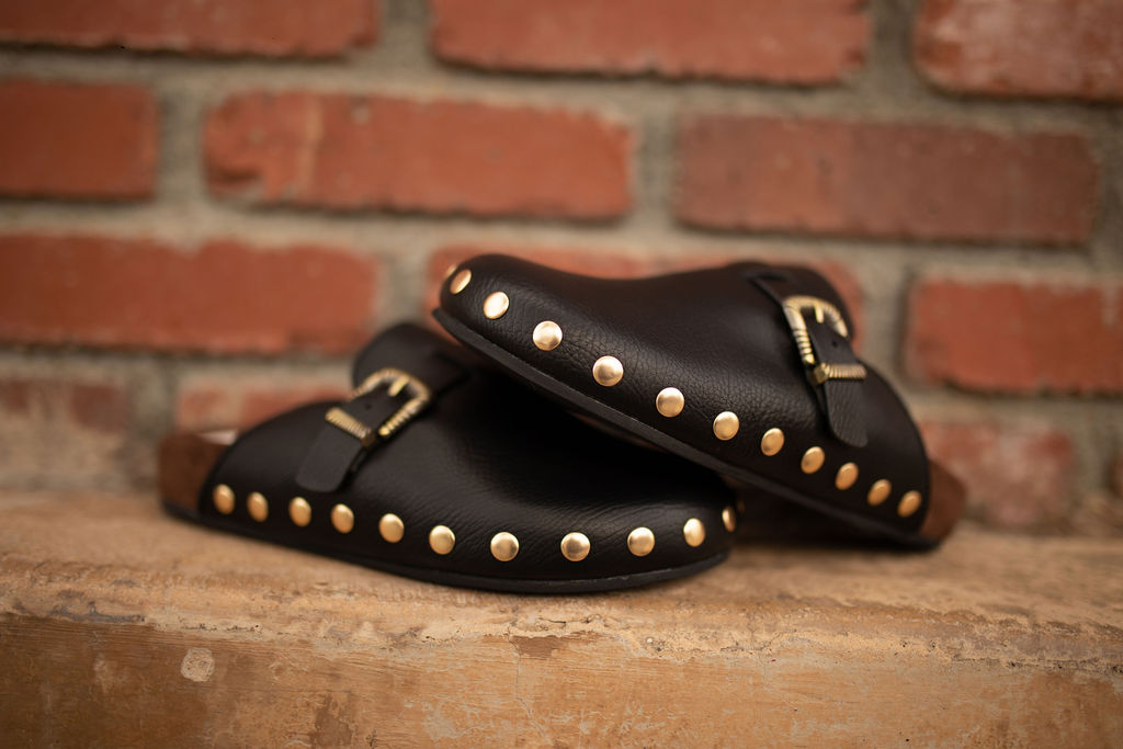Black leather clogs with gold studs and a gold buckle strap displayed against a rustic brick background.