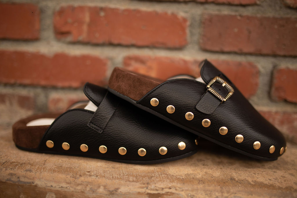 Black leather clogs with gold studs and a gold buckle strap displayed against a rustic brick background.