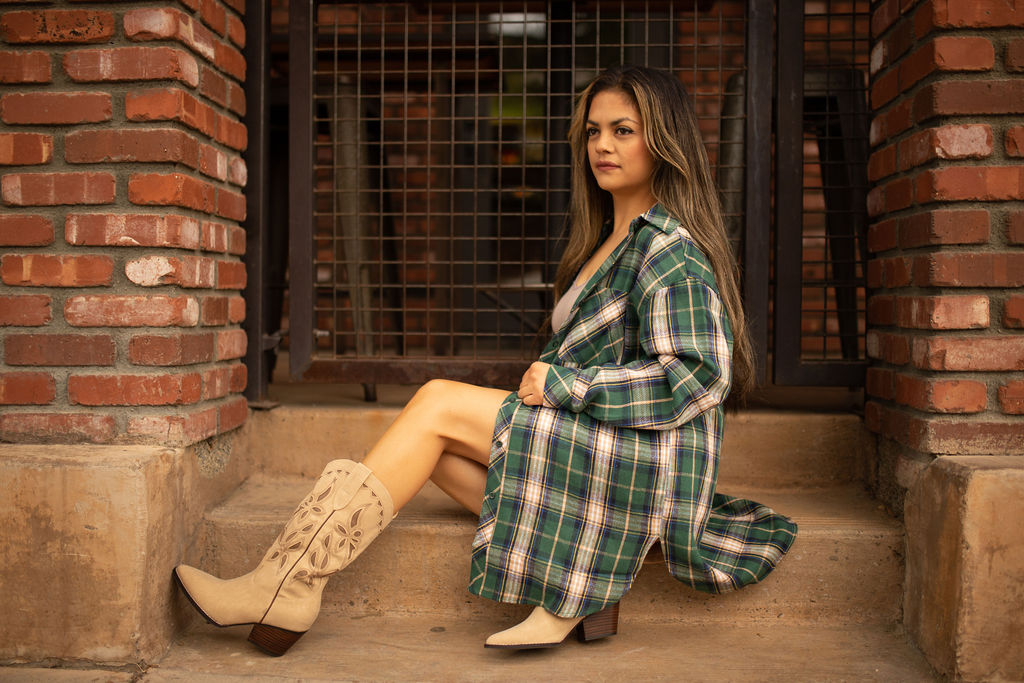Woman wearing a forest green and brown plaid oversized tunic top while sitting on outdoor steps, styled with tan western boots.