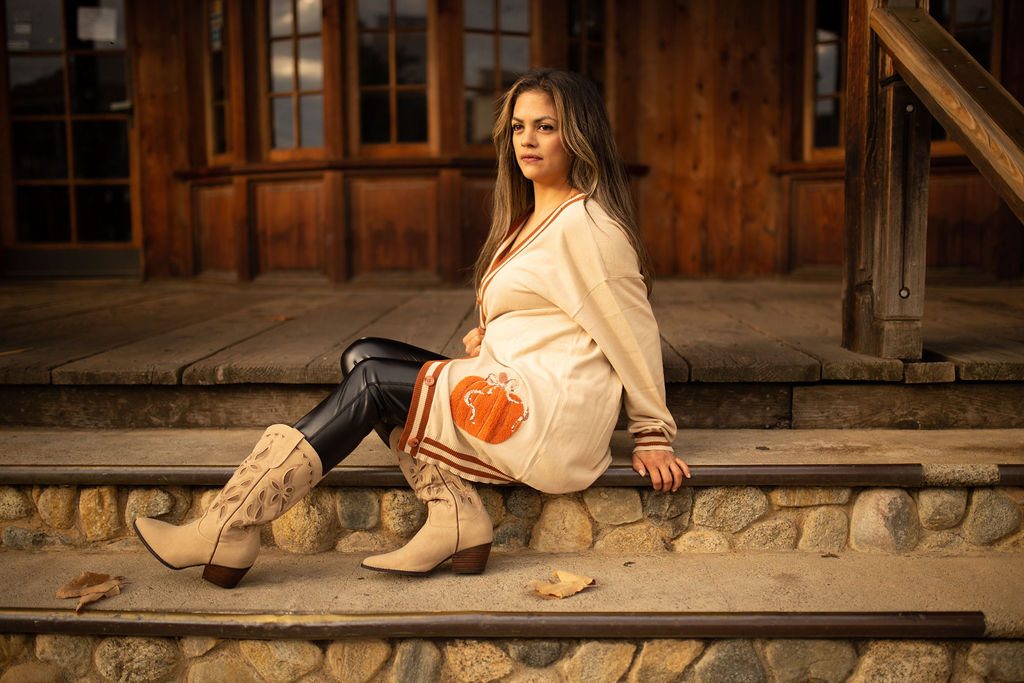 Cream cardigan with striped trim and an embroidered orange pumpkin patch on the lower side, shown in warm outdoor lighting.