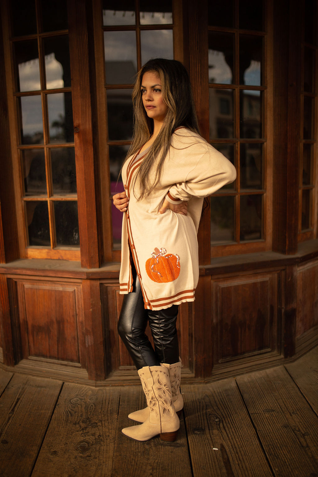 Cream cardigan with striped trim and an embroidered orange pumpkin patch on the lower side, shown in warm outdoor lighting.