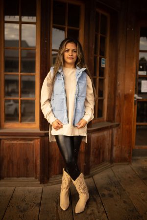 Light-wash denim puffer vest with quilted texture, high collar, and front pockets styled over a neutral long sleeve top.